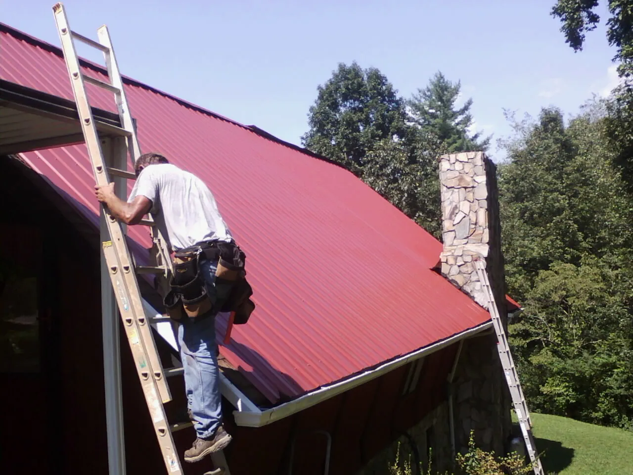 Roofer on a ladder beside a red metal roof and stone chimney — Soffit & Fascia Repair by Roofing Brothers in Wyoming